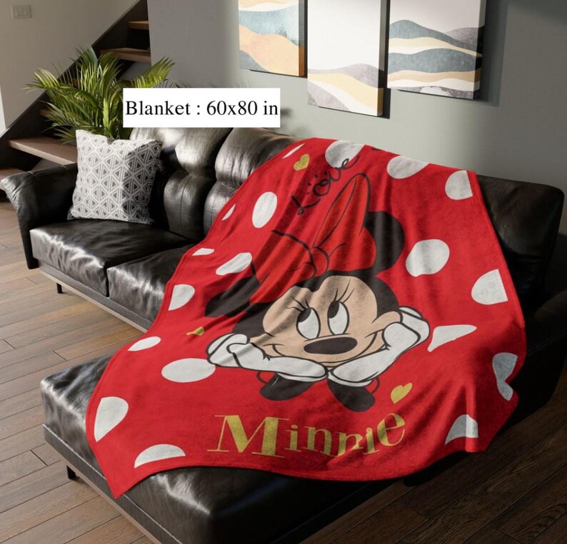 May include: A red blanket with white polka dots and a Minnie Mouse graphic. The blanket, measuring 60x80 inches, features Minnie Mouse with a red bow and the name "Minnie" in gold lettering. The blanket is draped over a black leather couch.