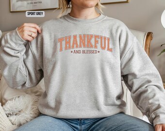 May include: A light grey sweatshirt with the words "THANKFUL AND BLESSED" in orange block letters. The sweatshirt is a crew neck style and is made of a soft material. The text is centered on the chest.