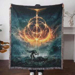 Könnte beinhalten: Eine gewebte Wurmdecke mit einer Fantasy-Landschaft mit einem leuchtenden, kreisförmigen Design in Orange- und Goldtönen. Eine Silhouette kniet mit einem Schwert im Vordergrund. Die Decke hat einen Fransenrand.