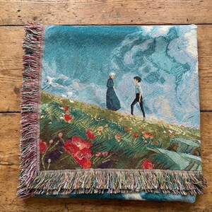 May include: Woven throw blanket featuring a pastoral scene with two figures on a hillside with red flowers. The blanket has a teal background with a sky and cloud design. The edges are finished with a fringed border.