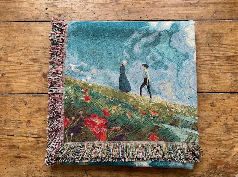 May include: Woven throw blanket featuring a pastoral scene with two figures on a hillside with red flowers. The blanket has a teal background with a sky and cloud design. The edges are finished with a fringed border.