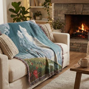 May include: A cozy living room scene with a cream-colored sofa draped with a blue and green landscape tapestry throw blanket. Two knitted throw pillows and a lit fireplace add warmth. A wooden coffee table holds a mug and candle.