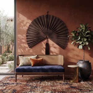 May include: A dark brown pleated wall fan with a central tassel hanging above a wooden settee featuring a cane backrest and a navy blue velvet seat cushion. The settee is adorned with a patterned beige pillow and a solid rust-colored velvet pillow.