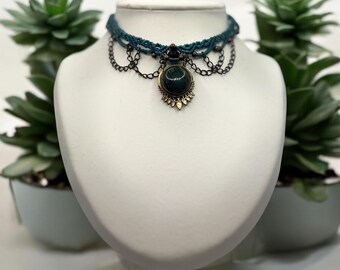 May include: Teal and black choker necklace with a large round green gemstone pendant. The pendant has a gold-toned setting with a decorative design.