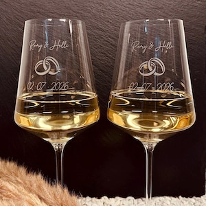 Può includere: Due calici da vino in vetro trasparente riempiti di vino bianco. Ogni calice è inciso con i nomi Romy & Helli, un'illustrazione di due fedi nuziali intrecciate e la data 02.07.2026 in un sottile carattere bianco corsivo e stampato.