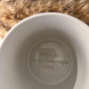 Könnte beinhalten: Eine weiße Keramiktasse mit dem deutschen Text "Willst du meine Trauzeugin sein?" im Inneren. Die Tasse hat eine gesprenkelte Textur und einen gebogenen Henkel. Getrocknetes Ziergras befindet sich im Hintergrund.