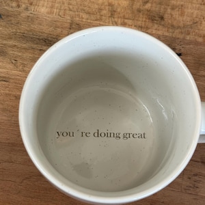 Può includere: Una tazza in ceramica bianca con un motivo maculato. L'interno della tazza presenta la frase "you're doing great" in grigio. La tazza ha una forma arrotondata e un manico.