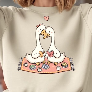 May include: A light beige sweatshirt featuring a whimsical illustration of two white geese with books, sitting on a pink rug. A pink heart floats above them. The geese are surrounded by small books and mugs.