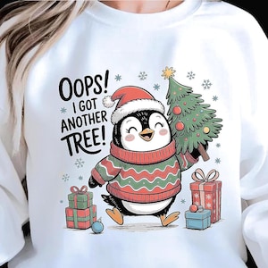 Puede incluir: Sudadera blanca con un pingüino de dibujos animados con un gorro de Papá Noel y un suéter rojo y verde, sosteniendo un árbol de Navidad. El texto "OOPS! I GOT ANOTHER TREE!" está encima del pingüino. También hay regalos y copos de nieve.