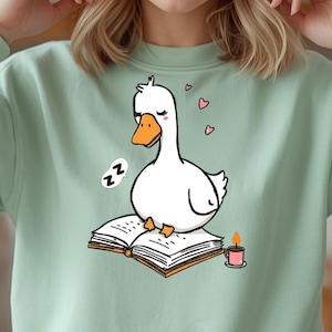 May include: A pale green sweatshirt with a cartoon goose sleeping on a book. The goose is white with an orange beak and feet. A small candle and two hearts are also depicted. The goose has closed eyes and a speech bubble with "Zz" in it.