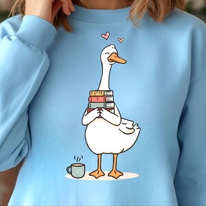 May include: Light blue sweatshirt featuring a cartoon goose holding a stack of books. The goose has an orange beak and feet, with two pink hearts above its head. A small coffee cup is to the left of the goose.