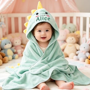 Peut inclure: Un bébé enveloppé dans une serviette à capuche vert clair avec un motif de dinosaure. La capuche a des pointes jaunes et le nom "Alice" brodé dessus. Le bébé est entouré de peluches dans un berceau.