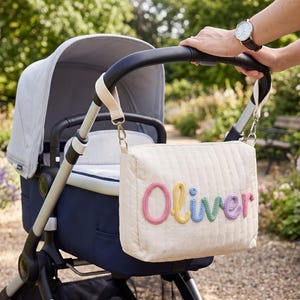 May include: A cream-colored quilted stroller organizer bag featuring the name Oliver in colorful, textured rope lettering. The bag is attached to the black handlebar of a navy blue and light gray stroller with two adjustable straps and metal clips.