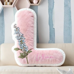 May include: A pink, plush letter "L" shaped cushion with a white border. The cushion features an embroidered floral design with blue and green accents. The name "Lillian" is written in pink script.