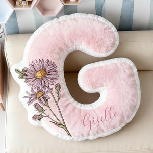 Può includere: Un cuscino a forma di lettera "G" rosa e soffice, con bordo bianco e un design floreale. Il cuscino presenta un'illustrazione ad acquerello di fiori viola e il nome "Giselle" in corsivo. Il cuscino è su un divano color crema.
