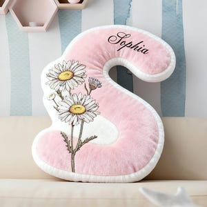 Peut inclure: Un coussin en forme de lettre "S" rose et moelleux avec une bordure blanche, orné d'un motif floral de marguerites et du nom "Sophia" en écriture cursive. Le coussin est sur un canapé beige contre un mur rayé.