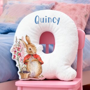 May include: A white, plush letter "Q" pillow with the name "Quincy" in blue script. The pillow features a watercolor illustration of a rabbit in a red cape, holding a basket of berries. The pillow is on a pink chair.