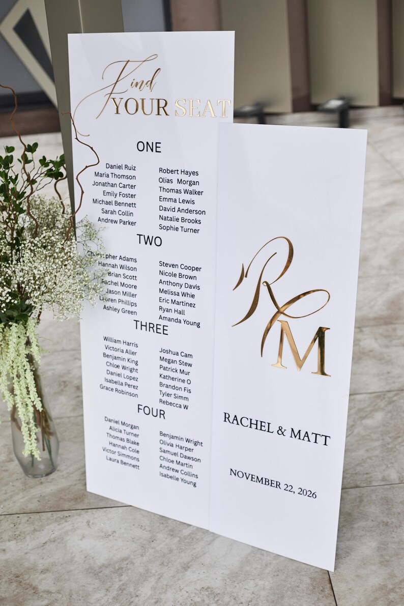 May include: A white wedding seating chart with gold lettering. The chart is divided into sections labeled "ONE," "TWO," "THREE," and "FOUR," listing names. The right side features the names "Rachel & Matt" and the date "November 22, 2026."