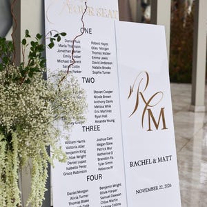 May include: White wedding seating chart with gold lettering. The chart is divided into four sections, each listing names under numbers. The top reads "Find Your Seat". The right side has the names "Rachel & Matt" and the date.