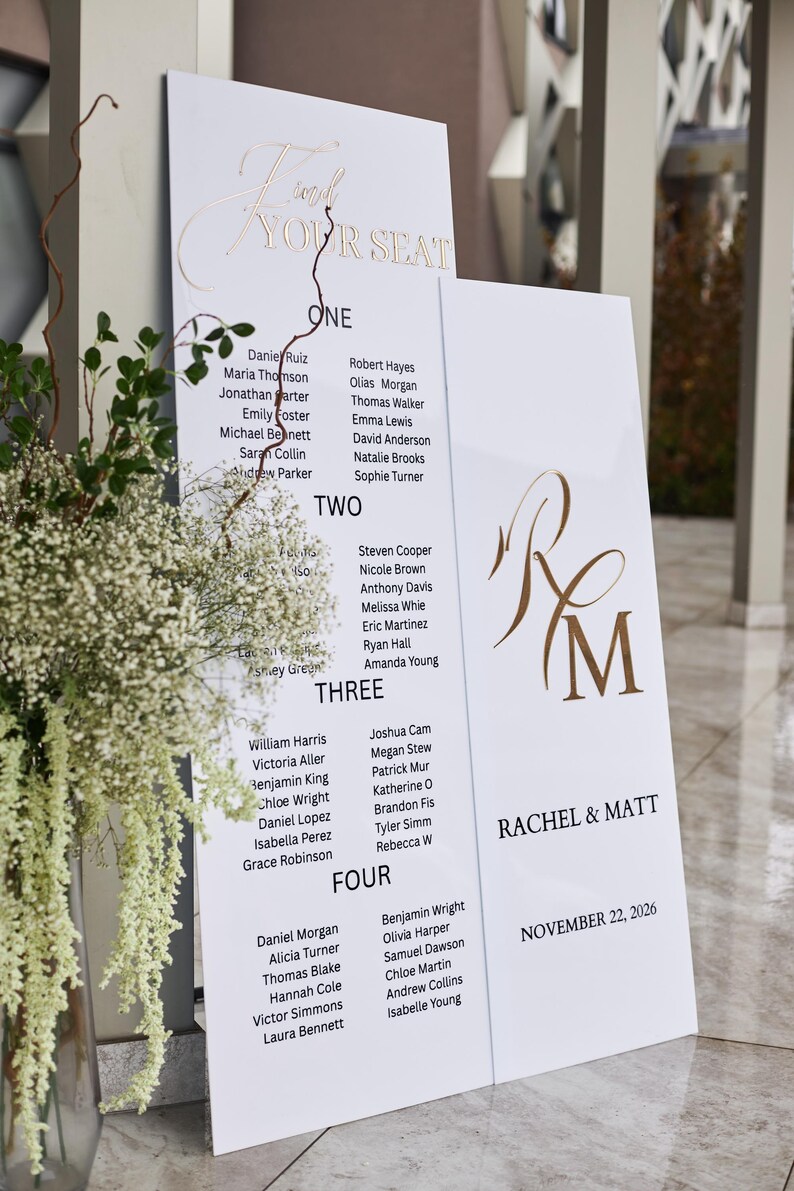 May include: White wedding seating chart with gold lettering. The chart is divided into four sections, each listing names under numbers. The top reads "Find Your Seat". The right side has the names "Rachel & Matt" and the date.