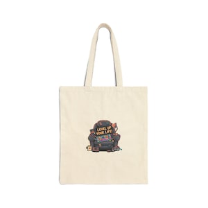 May include: Natural canvas tote bag with a vibrant graphic of a gaming chair adorned with gaming equipment. The text "LEVEL UP YOUR LIFE!" is displayed on a sign above the chair. The bag has two handles.
