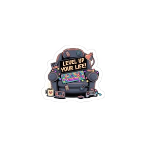May include: A sticker featuring a cartoon armchair with a keyboard, game controllers, and a coffee mug. The chair has a screen that reads "LEVEL UP YOUR LIFE!". A small lamp and other gaming accessories are also present.