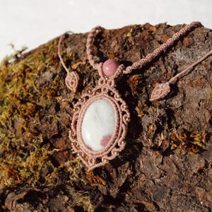 Puede incluir: Un collar de macramé hecho a mano con una gran piedra ovalada en blanco y rosa. La piedra está engastada en un diseño de macramé marrón claro detallado, con pequeñas cuentas doradas. Una esfera rosa y detalles de macramé en forma de hoja completan el collar.