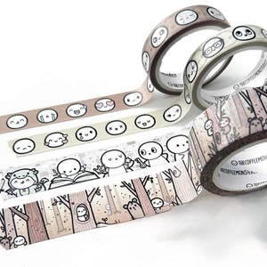 May include: Several rolls of decorative washi tape with various designs. One roll features a woodland scene with bears and trees. Other rolls display repeating patterns of cartoon faces and expressions. The tape is in shades of brown, beige, and white.