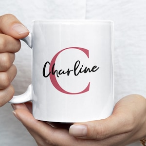 Peut inclure: Tasse en céramique blanche avec le nom "Charline" en écriture noire et un grand "C" rose. La tasse est tenue par une personne portant une bague et un bracelet dorés. Un petit logo est visible en haut à droite.