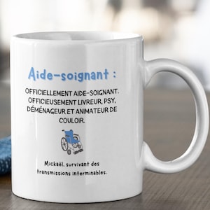 May include: White ceramic mug with a large handle. The mug features blue text that reads "Aide-soignant:" and additional text below. A small blue wheelchair graphic is also present. The mug is on a wooden surface.