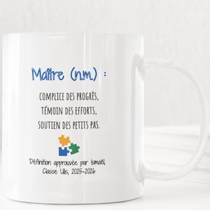Pode incluir: Uma caneca de cerâmica branca com uma alça grande. A caneca apresenta texto azul que diz "Maître (nm): COMPLICE DES PROGRÈS, TÉMOIN DES EFFORTS, SOUTIEN DES PETITS PAS." Abaixo, um gráfico de peças de quebra-cabeça. A caneca também inclui o texto "Définition approuvée par Ismaël Classe Ulis, 2025-2026."