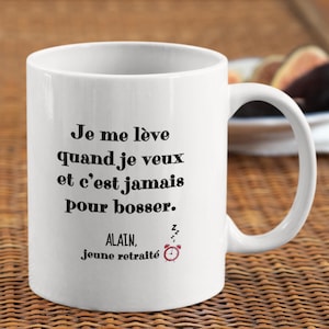 May include: White ceramic mug with black text: "Je me lève quand je veux et c'est jamais pour bosser. ALAIN, jeune retraité." A small alarm clock graphic is next to the text. The mug has a curved handle.