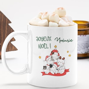Op de afbeelding: Een witte keramische mok gevuld met marshmallows, voorzien van een afbeelding van zes cartoonkatten met kerstmutsen op, gestapeld in een piramide. De tekst "JOYEUX NOËL !" en de naam "Noémie" zijn in het groen erboven gedrukt.