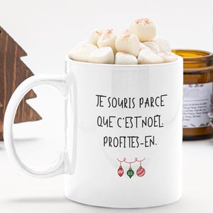 Puede incluir: Una taza de cerámica blanca llena de malvaviscos, con el texto en negro 'JE SOURIS PARCE QUE C'EST NOEL. PROFITES-EN.' y una pequeña ilustración de tres adornos navideños coloridos colgando en la parte inferior.