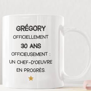 May include: White ceramic mug with the name "GRÉGORY" and the text "OFFICIELLEMENT 30 ANS OFFICIEUSEMENT : UN CHEF-D'OEUVRE EN PROGRÈS" printed in black. A gold star is at the bottom. The mug has a curved handle.
