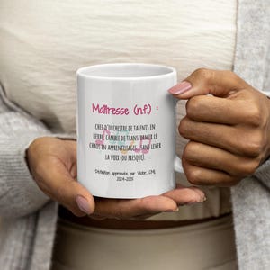 Pode incluir: Uma caneca de cer&acirc;mica branca segurada por uma pessoa, com o texto "Ma&icirc;tresse (n.f.): CHEF D'ORCHESTRE DE TALENTS EN HERBE, CAPABLE DE TRANSFORMER LE CHAOS EN APPRENTISSAGES... SANS LEVER LA VOIX (OU PRESQUE)." em rosa e preto. A caneca tamb&eacute;m inclui o texto "D&eacute;finition approuv&eacute;e par Victor, CML 2024-2025."