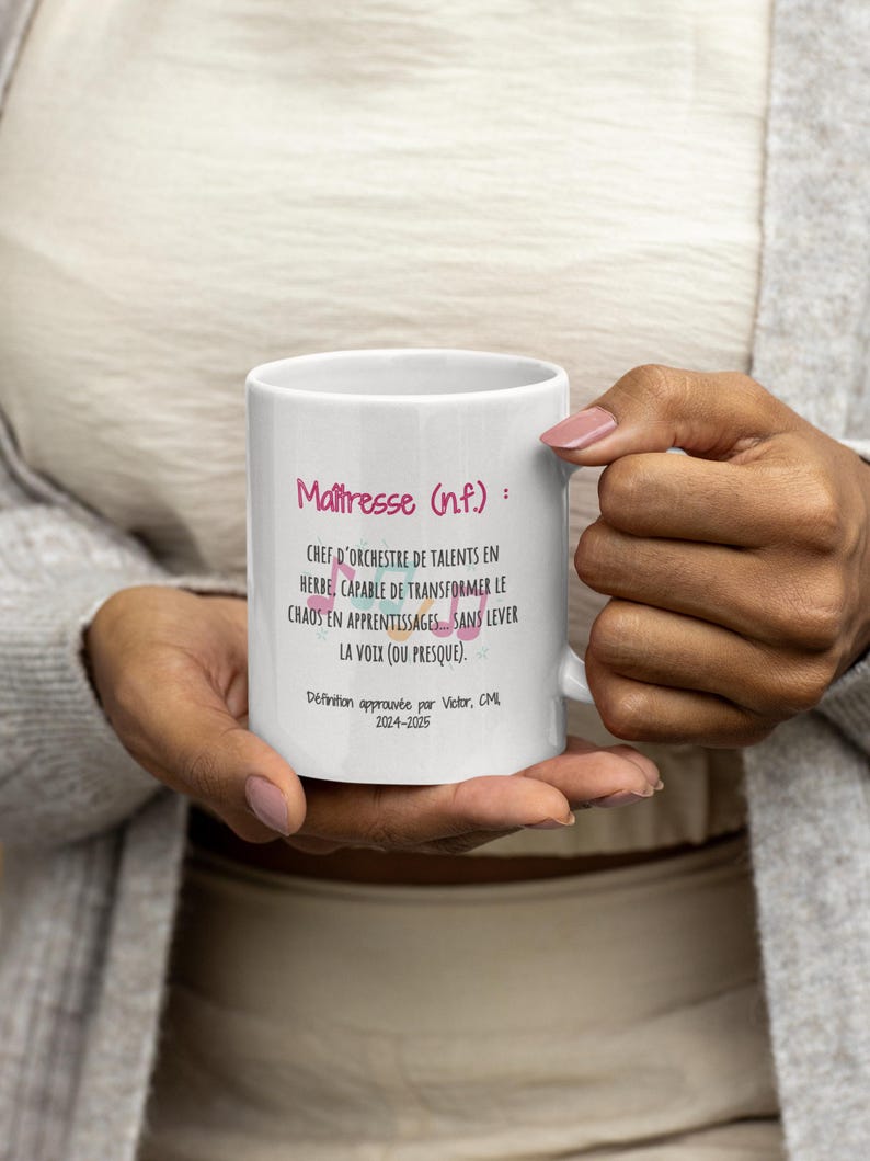 Pode incluir: Uma caneca de cer&acirc;mica branca segurada por uma pessoa, com o texto "Ma&icirc;tresse (n.f.): CHEF D'ORCHESTRE DE TALENTS EN HERBE, CAPABLE DE TRANSFORMER LE CHAOS EN APPRENTISSAGES... SANS LEVER LA VOIX (OU PRESQUE)." em rosa e preto. A caneca tamb&eacute;m inclui o texto "D&eacute;finition approuv&eacute;e par Victor, CML 2024-2025."