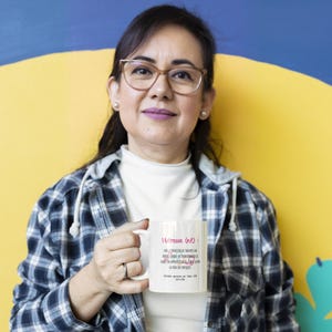 Pode incluir: Uma caneca de cer&acirc;mica branca com o texto "Ma&icirc;tresse (n.f.)" em rosa, juntamente com texto adicional em franc&ecirc;s. A caneca &eacute; segurada por uma pessoa que usa uma camisa xadrez e &oacute;culos. O fundo &eacute; amarelo e azul.