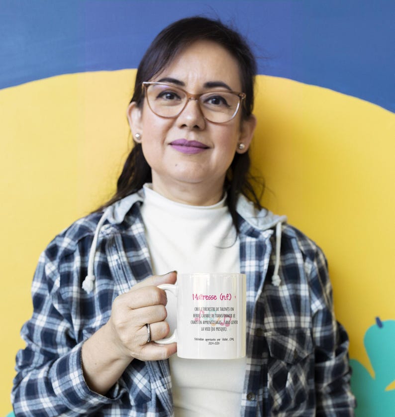 Pode incluir: Uma caneca de cer&acirc;mica branca com o texto "Ma&icirc;tresse (n.f.)" em rosa, juntamente com texto adicional em franc&ecirc;s. A caneca &eacute; segurada por uma pessoa que usa uma camisa xadrez e &oacute;culos. O fundo &eacute; amarelo e azul.