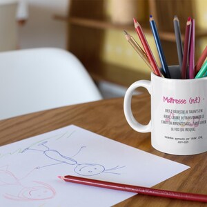 Pode incluir: Uma caneca de cer&acirc;mica branca com l&aacute;pis coloridos, com o texto "Ma&icirc;tresse (nf)" e outro texto em franc&ecirc;s. Um desenho e um l&aacute;pis vermelho est&atilde;o sobre a mesa de madeira. Um item tem&aacute;tico para professores.