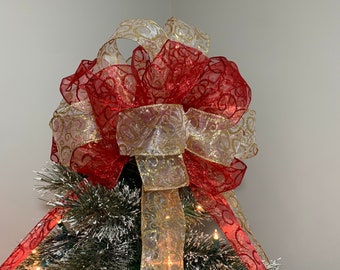 Könnte beinhalten: Ein roter und goldener Weihnachtsbaumspitzen-Bogen mit glitzernder Oberfläche. Die Schleife besteht aus zwei verschiedenen Arten von Band, einem mit einem roten und goldenen Wirbelmuster und einem mit einem goldenen Wirbelmuster. Die Schleife ist mit einem großen Knoten in der Mitte gebunden.