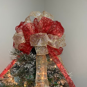 May include: A red and gold Christmas tree topper bow with a glittery finish. The bow is made of two different types of ribbon, one with a red and gold swirl pattern and the other with a gold swirl pattern. The bow is tied with a large knot in the center.