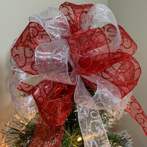 May include: A red and white Christmas tree topper bow with silver glitter. The bow is made of sheer ribbon with a swirl pattern.