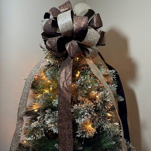 Könnte beinhalten: Ein brauner und goldener Glitzer-Christbaumspitzen-Bogen. Die Schleife besteht aus zwei verschiedenen Bändern, einem braunen und einem goldenen, und ist zu einer großen, flauschigen Schleife gebunden. Die Schleife ist perfekt, um jedem Weihnachtsbaum einen Hauch von Glanz zu verleihen.