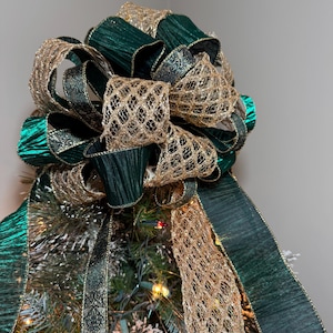 Op de afbeelding: Een kerstboomtopper met een grote strik gemaakt van smaragdgroene en gouden linten. De strik heeft een gelaagd ontwerp met getextureerde en open geweven elementen, wat een feestelijke en elegante uitstraling creëert. De boom is versierd met lichtjes en kunstsneeuw.