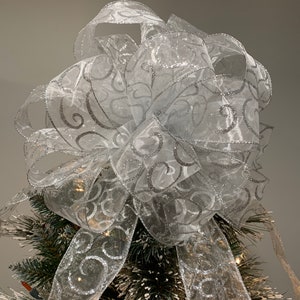 May include: A silver and white Christmas tree topper bow with a swirl design. The bow is made of sheer ribbon with silver glitter.