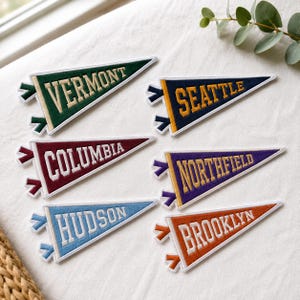 May include: Six triangular felt pennant patches with embroidered lettering. The patches feature the names Vermont in green, Seattle in navy, Columbia in maroon, Northfield in purple, Hudson in light blue, and Brooklyn in orange, all with white borders.
