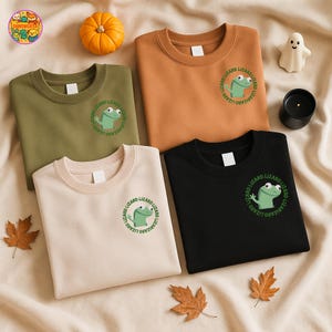 May include: Four crewneck sweatshirts in olive green, tan, brown, and black. Each features a green lizard graphic with the text "Lizard" in a circular design. Autumn-themed decor includes a pumpkin and leaves.