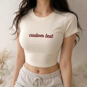 Könnte beinhalten: Ein cremefarbenes Kurzarm-Bauchfrei-Top mit Rundhalsausschnitt und dem Schriftzug "custom text", der in dunkelrotem Garn mittig auf der Brust eingestickt ist. Das Kleidungsstück ist körpernah geschnitten und aus glattem, einfarbigem Stoff.
