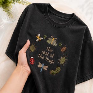 Pode incluir: Uma t-shirt preta de manga curta com um bordado circular de vários insetos, incluindo uma abelha, uma traça, uma joaninha, uma libélula e uma lagarta, rodeando o texto "the last of the bugs" em fio de cor castanho claro.